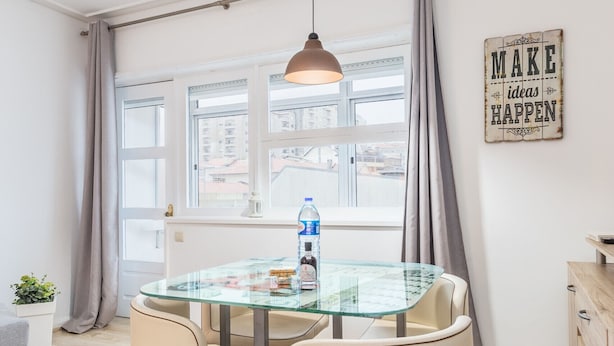 Un comedor moderno con una mesa de vidrio, dos sillas, una lámpara colgante y vistas de la ciudad a través de amplios ventanales.
