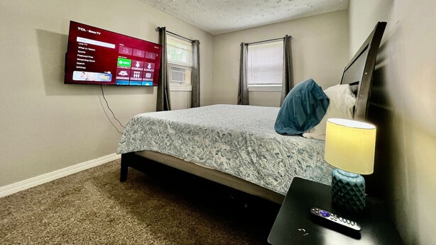 Un dormitorio con cama, televisor montado en la pared, mesita de noche con lámpara y ventana con cortinas.