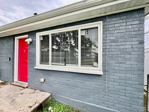 Ein Wohngebäude mit roter Tür, einem Fenster mit Rollläden und einer Fassade aus grauen Ziegeln.