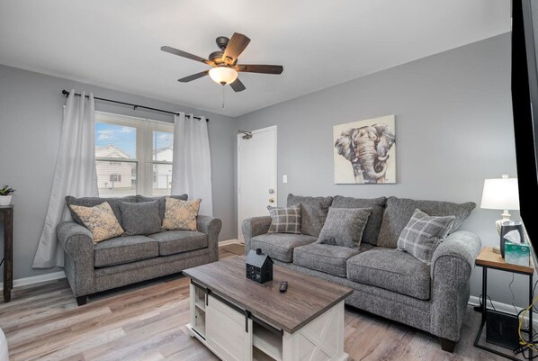 A living room with two grey sofas, a wooden coffee table, a ceiling fan, and a window with curtains.