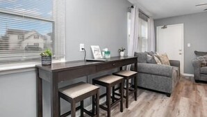 A modern living room with a grey sofa, a dark wooden desk with two stools, and a window with blinds. - 17 S First - Unit 6 · 17 s first street unit 6 (Trenton)