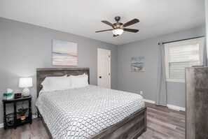 Un dormitorio con cama, mesita de noche, ventilador de techo, puerta y ventana con cortinas.
