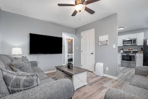 A modern living room with grey sofas, a flat-screen TV, a ceiling fan, and a kitchen area with white cabinets and appliances. - 17 S First - Unit 6 · 17 s first street unit 6 (Trenton)