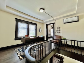 A room with a staircase, a desk with a chair, a window with blinds, and a wall-mounted air conditioning unit.