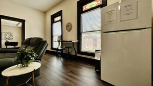 Un espacio habitable compacto con sofá, una mesita redonda, refrigerador y ventana con persianas.