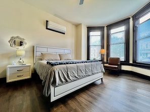 A bedroom with a bed, bedside tables, a lamp, a chair, and large windows with blinds.