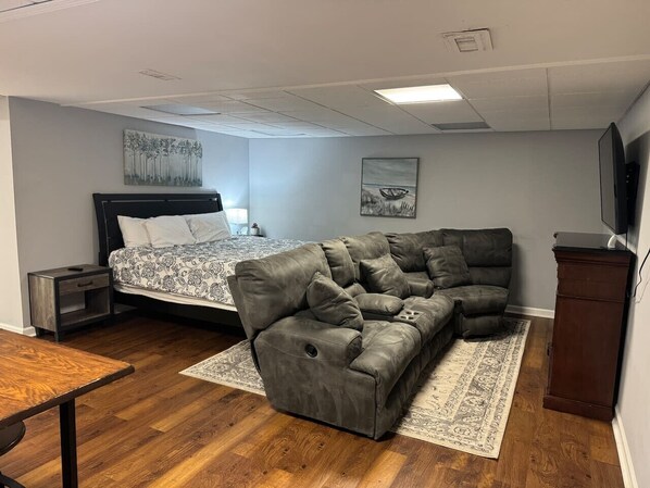 A bedroom with a bed, a gray sofa, a nightstand, a wooden table, and a TV.