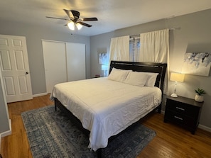 A bedroom with a bed, a nightstand, a lamp, a ceiling fan, a window with curtains, and a door.