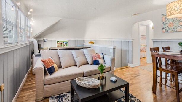 A cozy loft apartment with a high ceiling, a dining area with a wooden table and chairs, a sofa with cushions, and a kitchen in the background.