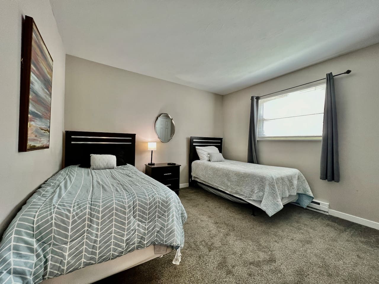A bedroom with two beds, a bedside table with a lamp, a window with curtains, and a wall-mounted mirror.