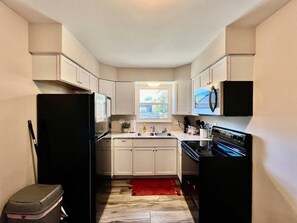 Une cuisine de taille réduite avec des meubles blancs, un réfrigérateur noir, une cuisinière et une fenêtre qui laisse entrer la lumière naturelle.