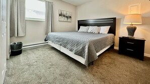 Une chambre à coucher comprenant un lit, une table de chevet, une lampe, une fenêtre avec des rideaux et une œuvre d’art murale.