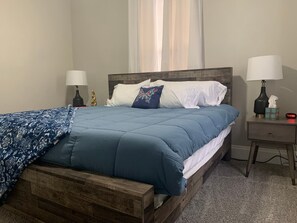 Un dormitorio con estructura de cama de madera, colcha azul, almohadas blancas y dos mesitas de noche con lámparas.