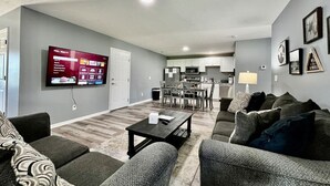 Una sala de estar con un televisor de pantalla plana, sofás grises, una mesa de centro y un comedor con sillas y una mesa.