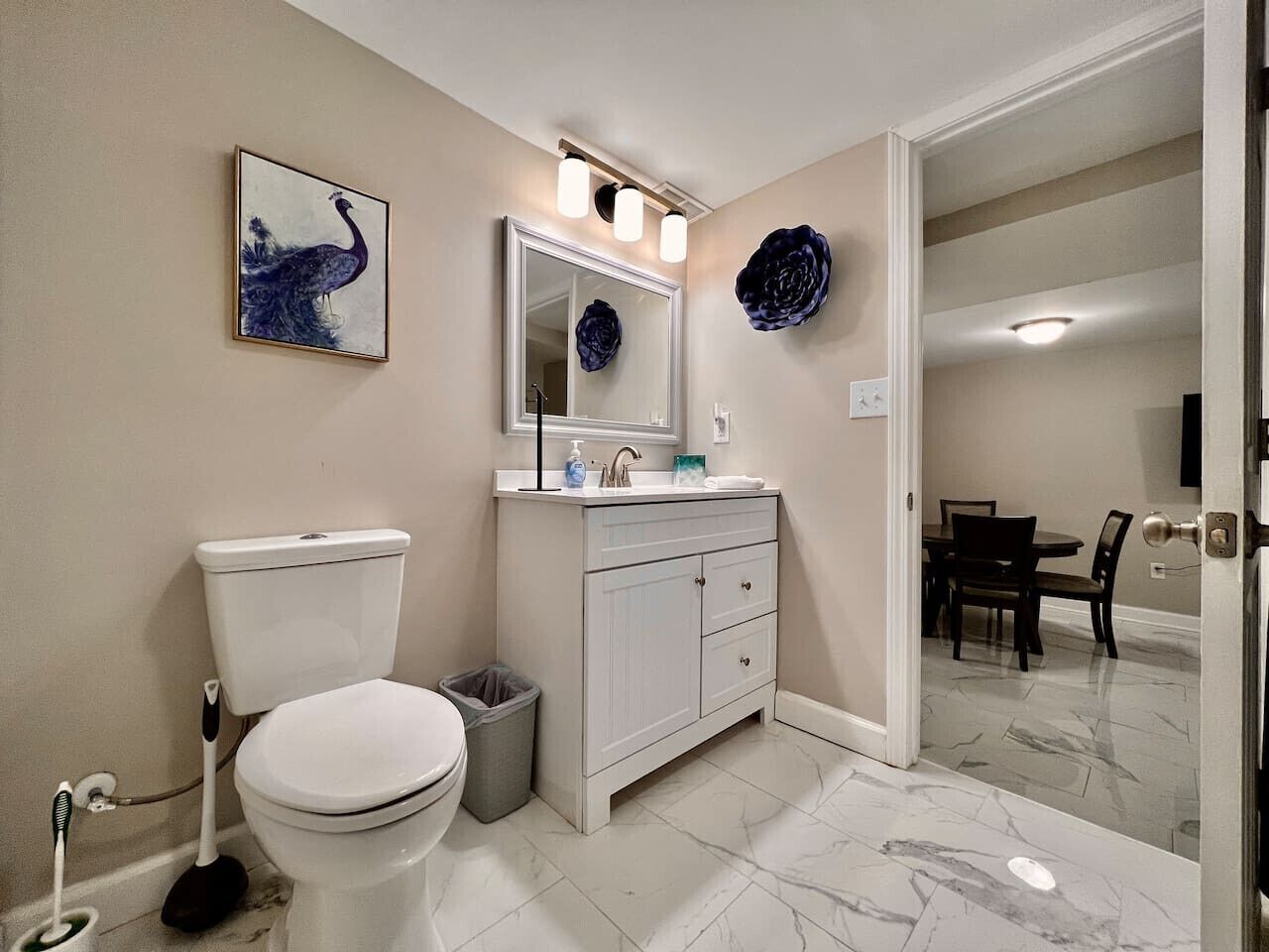 A bathroom with a toilet, sink, mirror, and a dining area visible through an open door.