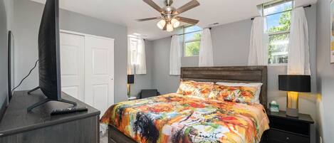 A bedroom with a bed, a TV on a stand, a ceiling fan, and a window with curtains.