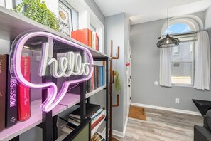 A modern living room with a bookshelf, a neon sign, a hanging plant, and a view of the outside. - 101 Crawford Street unit B · Funky Firehouse Renovated into Modern 2 BDR/1 Bath (Middletown)