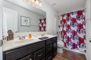 A bathroom with a double sink vanity, a toilet, a shower curtain with a floral pattern, and a small house-shaped decoration on the vanity.