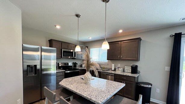 Una cocina moderna con electrodomésticos de acero inoxidable, una isla con encimera de granito, iluminación colgante y armarios de madera.
