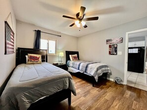 Ein Schlafzimmer mit zwei Betten, einem Deckenventilator, einer amerikanischen Flagge und einer durchgehenden Tür erkennbaren Küche.