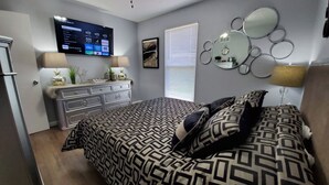 A bedroom with a bed, a TV mounted on the wall, a dresser, and a window with blinds.