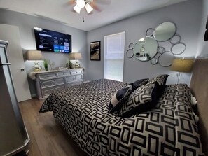 A bedroom with a bed, a TV mounted on the wall, a dresser, and a window with blinds.