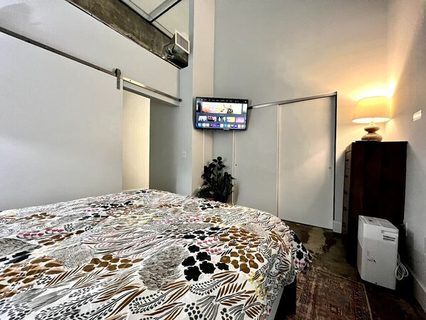 A bedroom with a bed, a television mounted on the wall, a lamp, and a small cabinet.