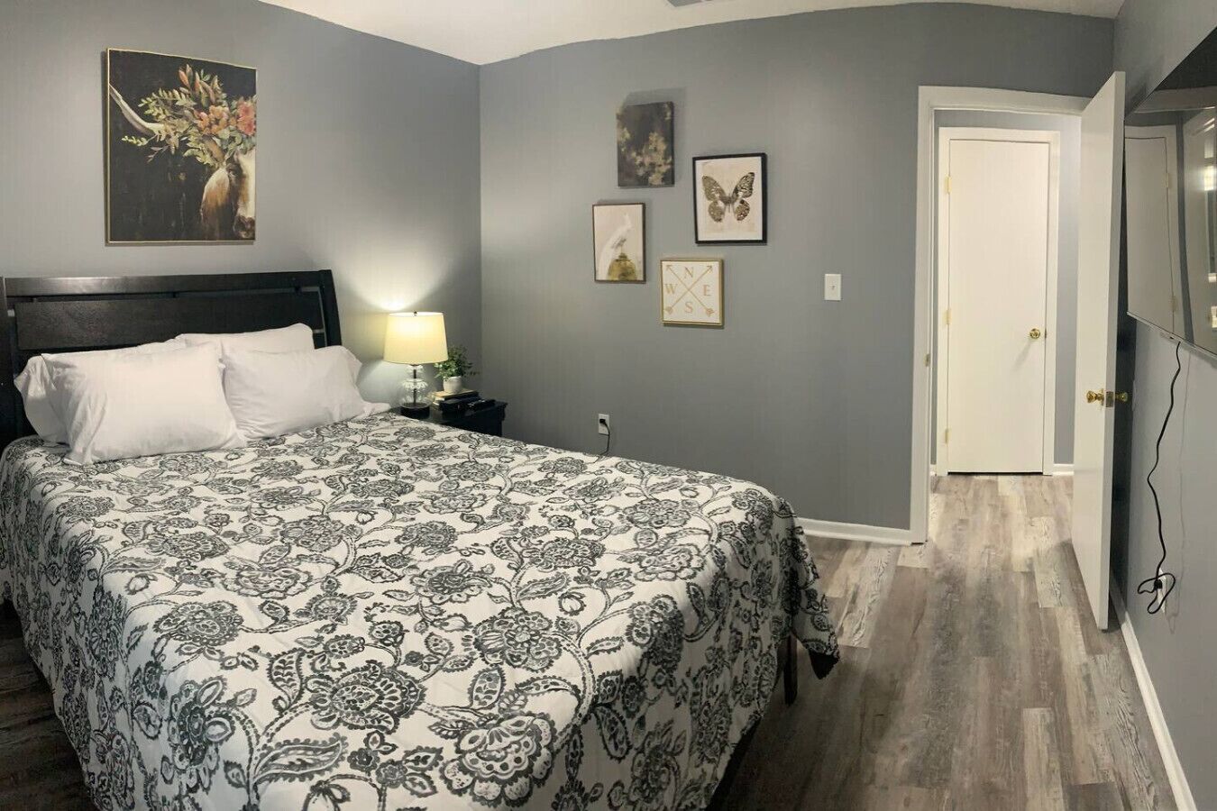A bedroom with a bed, a nightstand with a lamp, a door, and wall art.