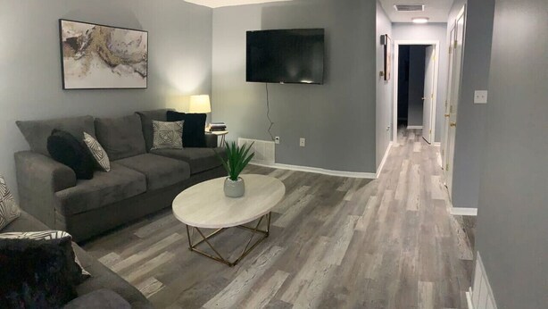 A living room with a grey couch, a round coffee table, a flat-screen TV mounted on the wall, and a hallway leading to another room. - 5145 Porsche · Newly Remodeled 2 BDRM Ranch- Attached Home (Liberty Township)
