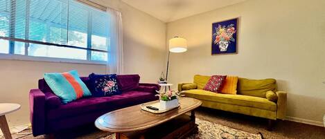 Una sala de estar con un sofá morado, un sofá amarillo, una mesa de centro de madera y un tapete estampado.