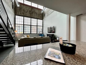 A spacious interior with a high ceiling, a large window, a sofa, a television, and a magazine on the floor. - 5101 Peters Cartridge · █Swanky loft at Historic Peters Cartridge Factory█ (Maineville)