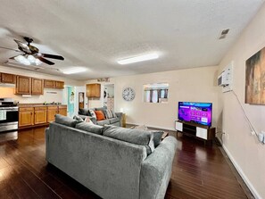 A living room with a grey sofa, a television, and a kitchen in the background. - 30 Royal #3 · Royal Treatment Studio near highway (Springboro)