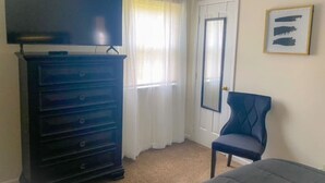 Een kamer met een grote donkerblauwe dressoir, een blauwe fauteuil, een bed, een deur en een raam met witte gordijnen.