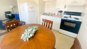 Een kleine keuken met witte kasten, een witte koelkast, een fornuis en een eettafel met stoelen.