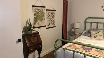 A bedroom with a green metal bed frame, a quilt, a small bedside table with a lamp, and two framed botanical illustrations on the wall.