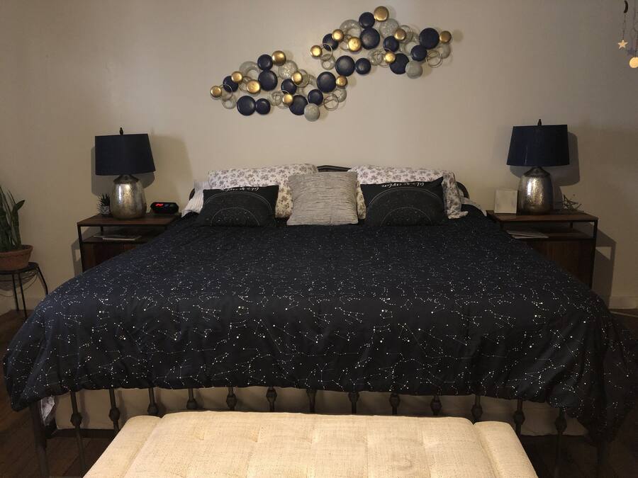 A bedroom with a large bed, two bedside tables with lamps, a nightstand, a potted plant, and a wall decoration.