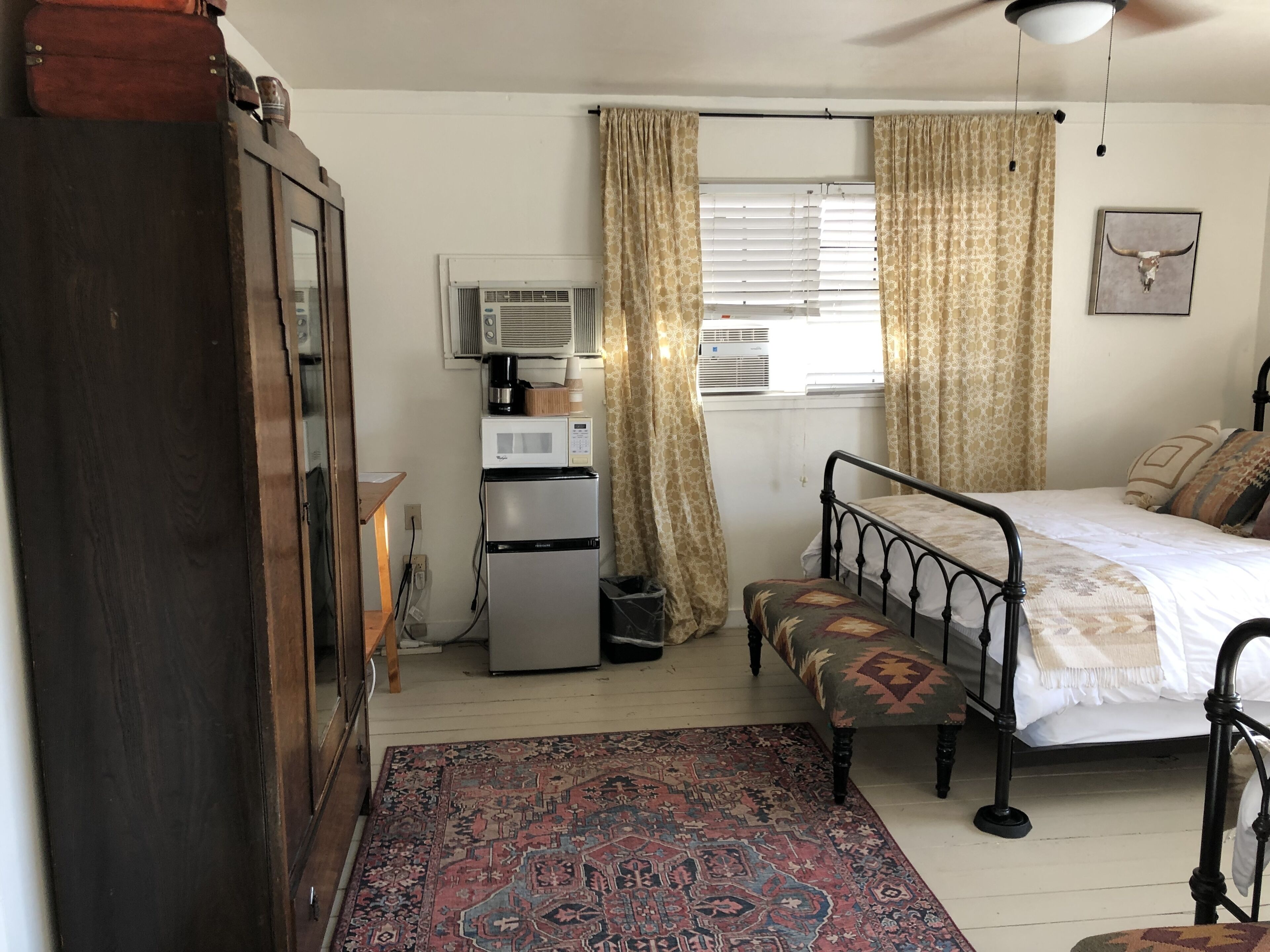 A bedroom with a bed, a bench, a wardrobe, a refrigerator, an air conditioner, and a patterned rug.