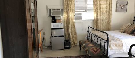 A bedroom with a bed, a bench, a wardrobe, a refrigerator, an air conditioner, and a patterned rug.