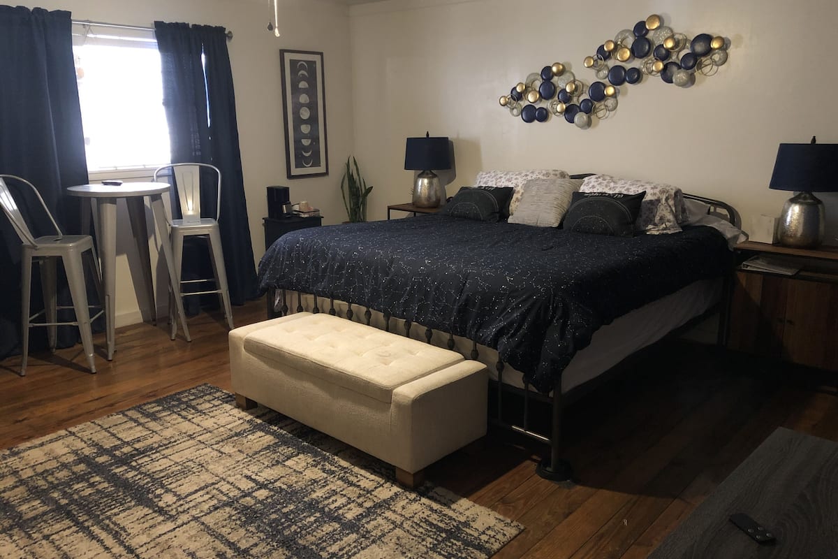 A bedroom with a bed, bedside tables, a small round table, two chairs, a bench, and a wall art piece.