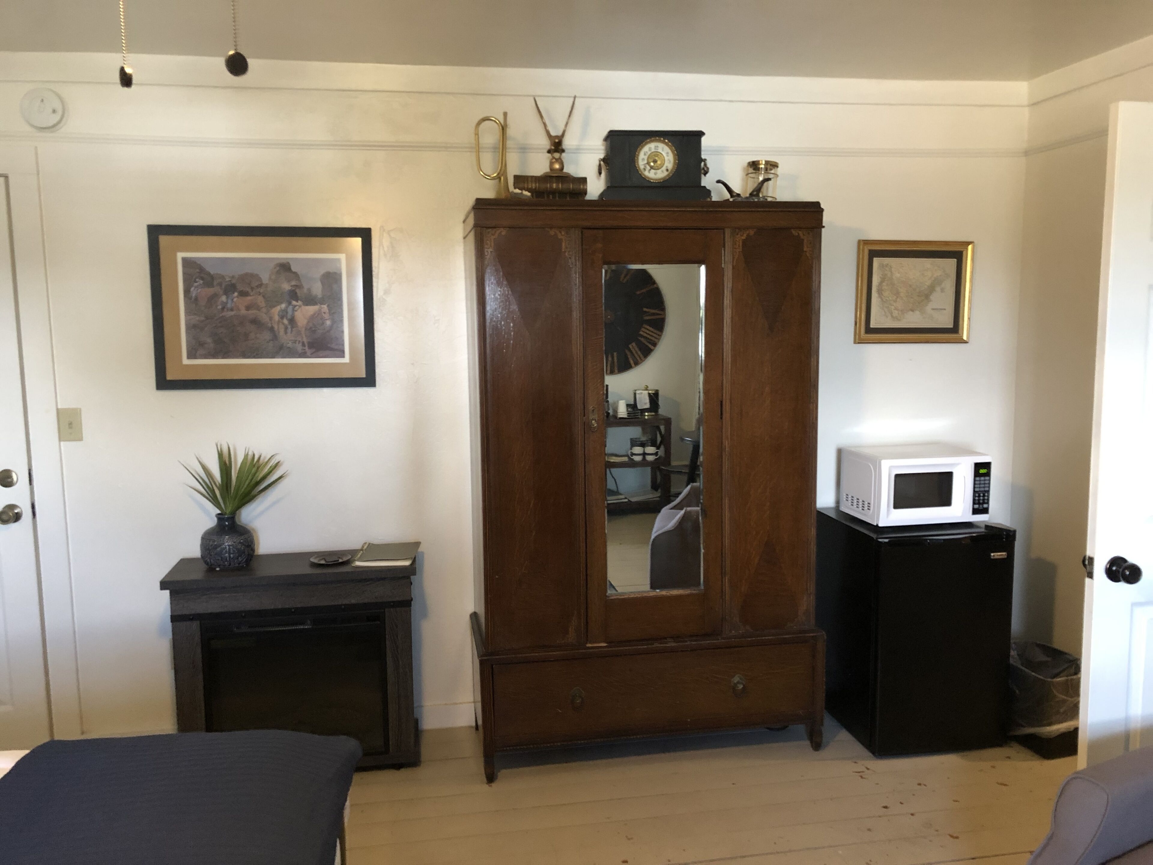 A room with a wooden wardrobe, a small desk with a plant, a framed picture, a clock, and a microwave.