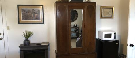 A room with a wooden wardrobe, a small desk with a plant, a framed picture, a clock, and a microwave.