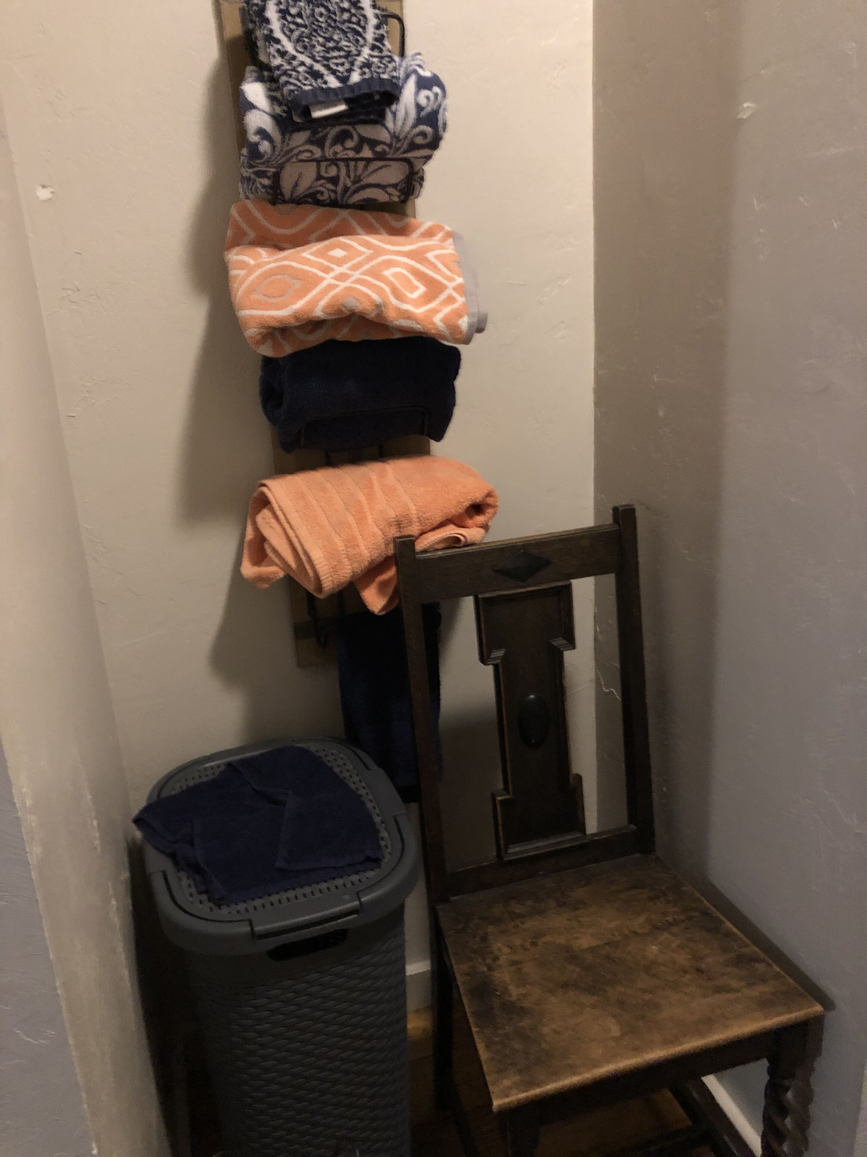 A small, dimly lit room with a wooden chair and a trash bin. A wall-mounted rack holds several folded towels.