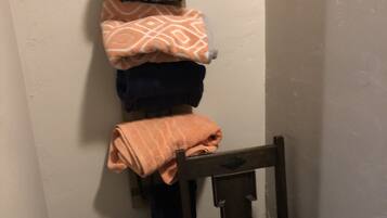 A small, dimly lit room with a wooden chair and a trash bin. A wall-mounted rack holds several folded towels.
