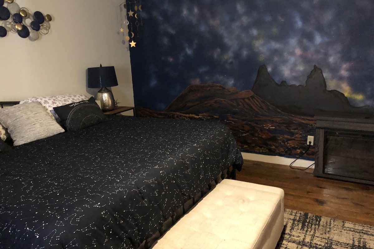 A bedroom with a bed, a night sky mural, a lamp, and a TV stand.