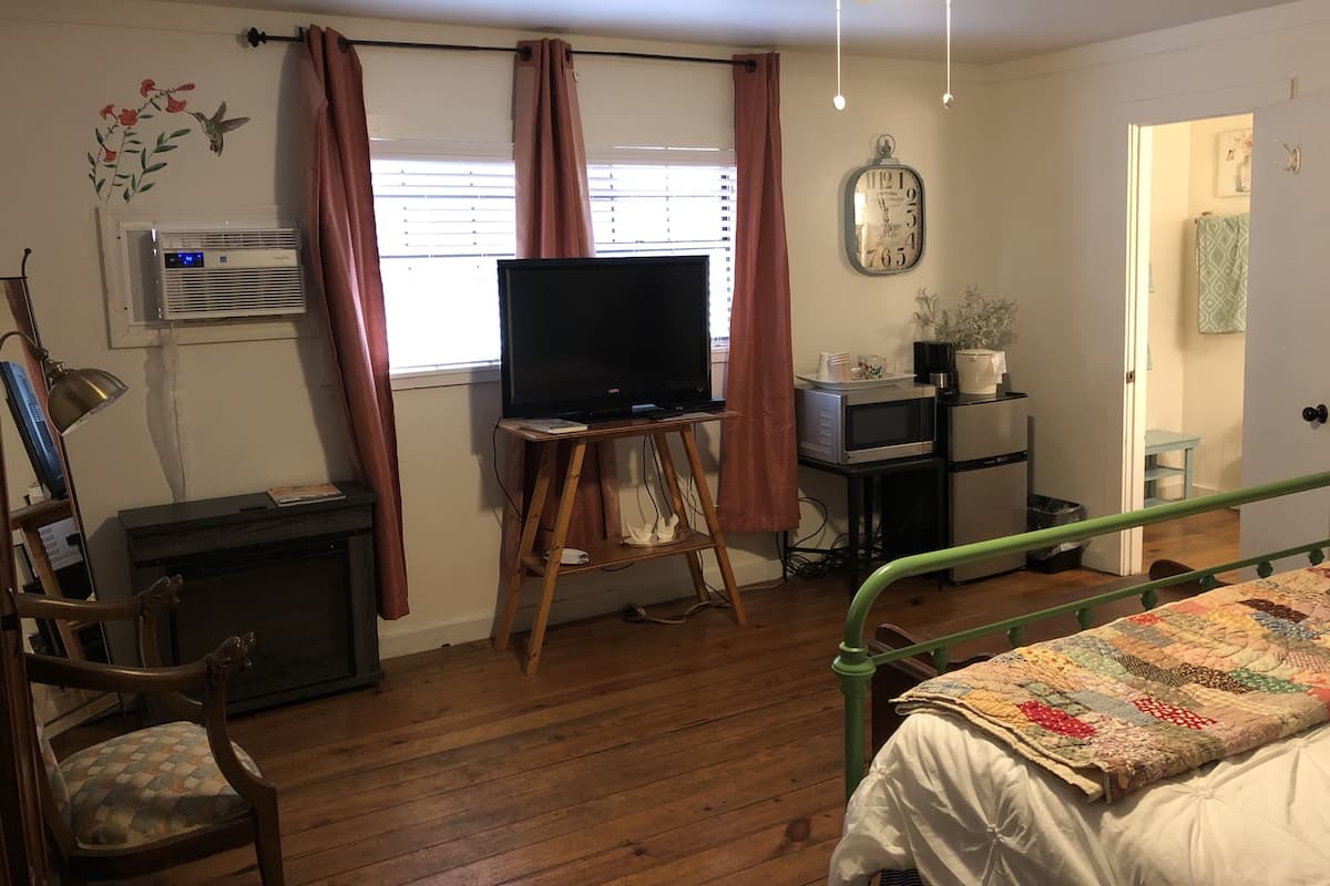 A bedroom with a wooden floor, a bed with a quilt, a television on a stand, a desk with a computer, a chair, and a window with curtains.