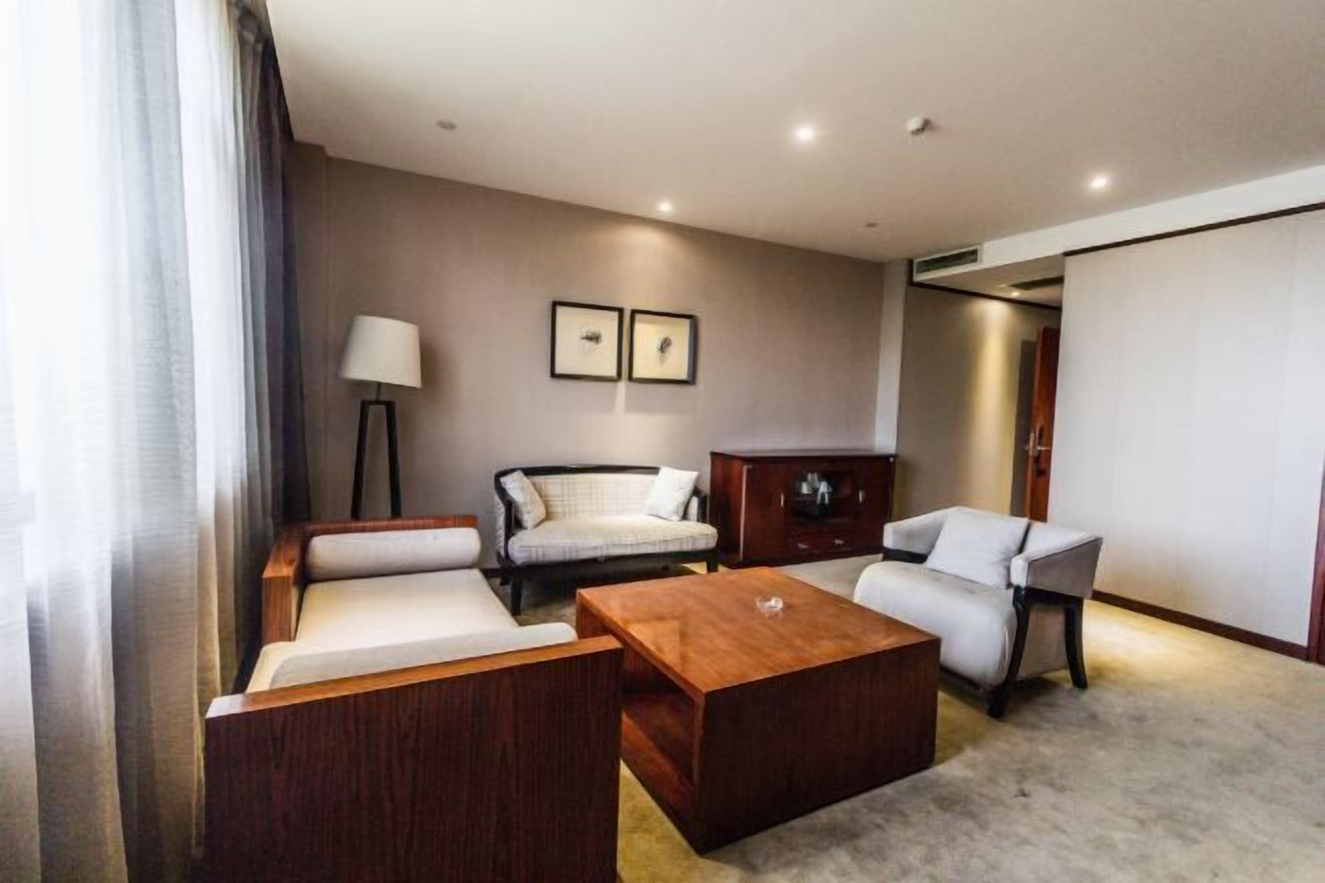 A modern hotel room with a sofa, armchair, and a wooden coffee table. There are two framed pictures on the wall, a lamp, and a wardrobe.