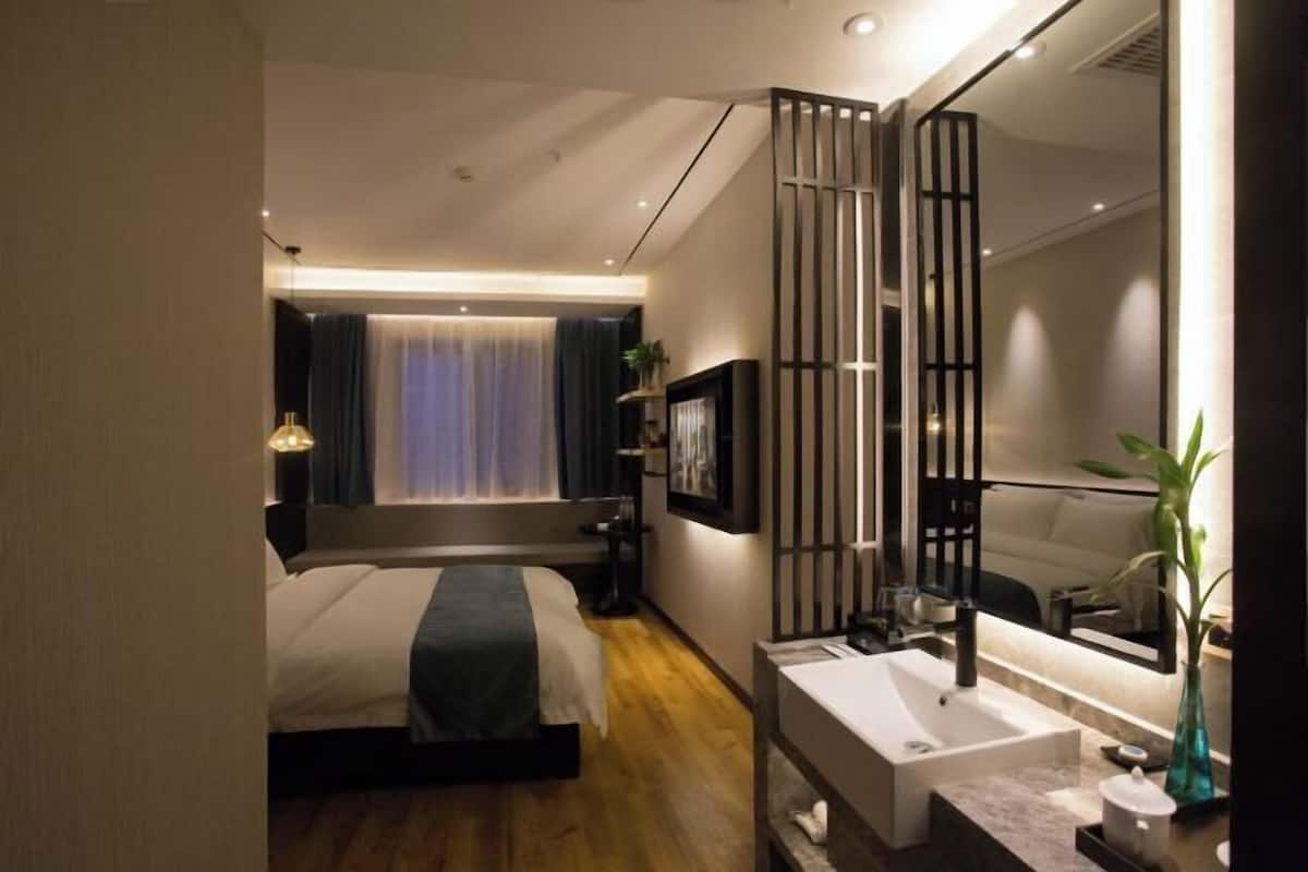 Una habitación de hotel moderna con cama, mesita de noche, tocador y un espejo grande, además de un baño con lavabo y espejo.