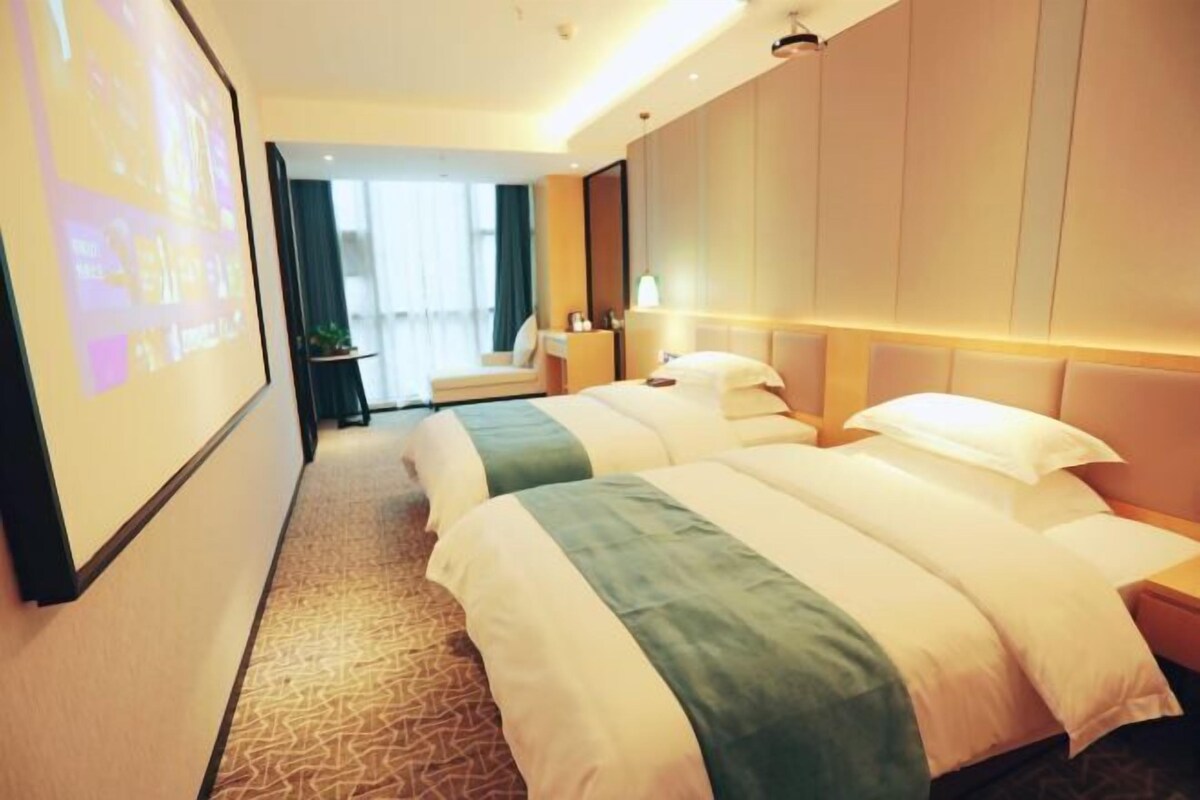 A hotel room with two beds, a large screen, a desk, and a view of the outdoors.
