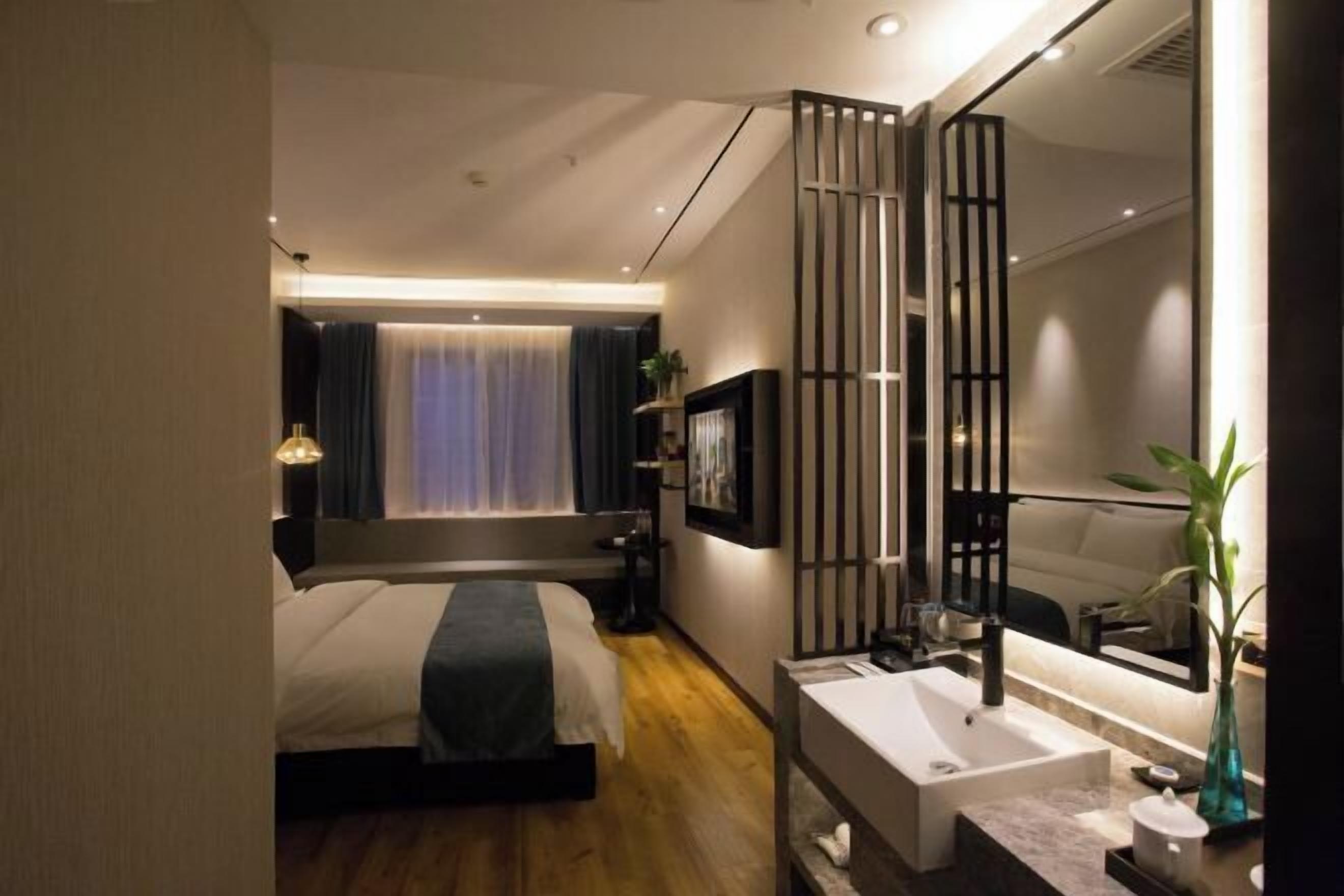 Una habitación de hotel moderna con una cama grande, una mesita de noche, un espejo, un baño con lavamanos y una ventana con cortinas.