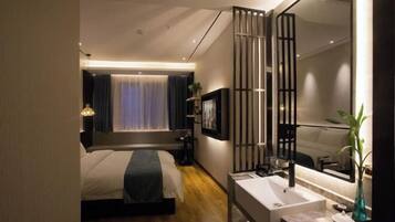A modern hotel room with a large bed, a bedside table, a mirror, a bathroom with a sink, and a window with curtains.